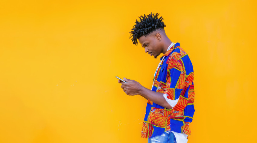 A man african print shirt stands his body the right looking at his phone against yellow background A man african print shirt stands his body the right looking at his phone against yellow background